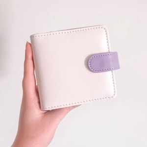 May include: A square-shaped bi-fold wallet in a soft cream color featuring a smooth finish and visible stitching along the edges. The wallet is secured with a small, rounded tab closure in a contrasting light purple color on the right side.