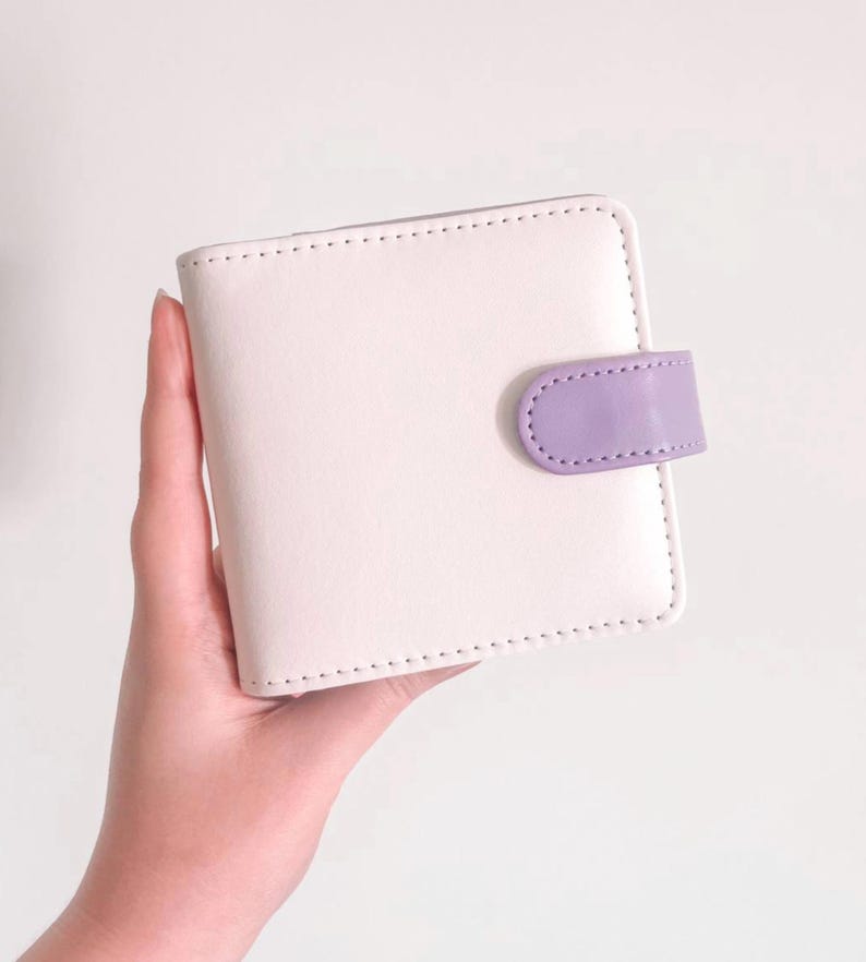 May include: A square-shaped bi-fold wallet in a soft cream color featuring a smooth finish and visible stitching along the edges. The wallet is secured with a small, rounded tab closure in a contrasting light purple color on the right side.