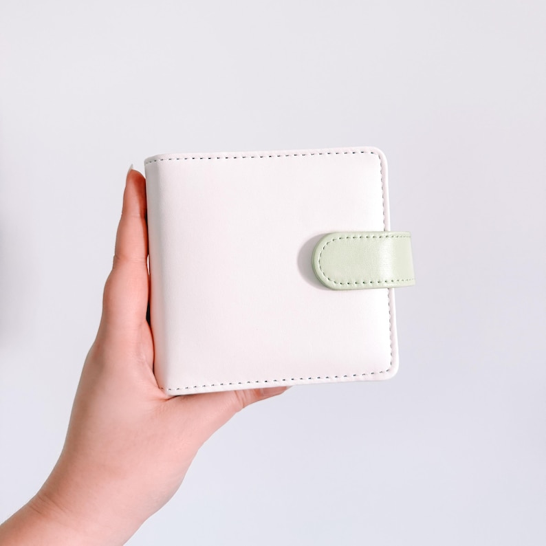 May include: A square white wallet featuring a smooth finish and a light green snap closure tab on the right side. The wallet has visible white stitching along the edges and a clean, simple design with a secure fastening mechanism for closure.