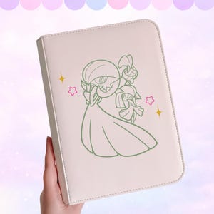 Può includere: Un raccoglitore color crema con un'illustrazione verde di tre personaggi dei cartoni animati e accenti di stelle e cuori. Il raccoglitore ha un bordo cucito e una superficie liscia.