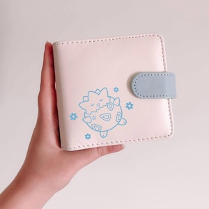 May include: A square, pale pink bi-fold wallet featuring a light blue snap closure tab. The front displays a light blue line art graphic of a small, round creature with triangular patterns and four small flower icons surrounding it.