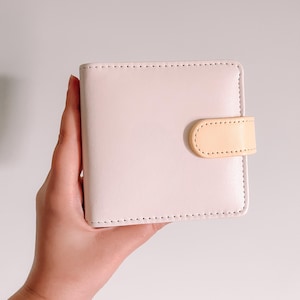 May include: A square-shaped, light pink wallet featuring a smooth, matte finish and rounded corners. The wallet is secured with a small, light peach-colored tab closure on the right side, which is attached with visible stitching along the edges.
