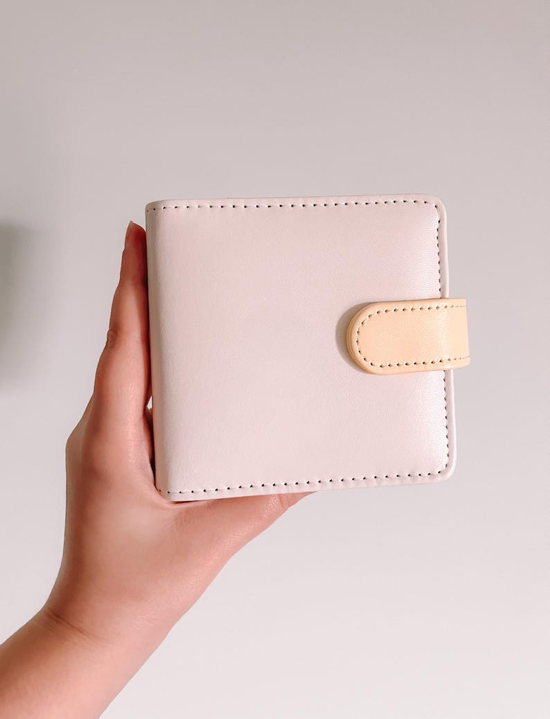 May include: A square-shaped, light pink wallet featuring a smooth, matte finish and rounded corners. The wallet is secured with a small, light peach-colored tab closure on the right side, which is attached with visible stitching along the edges.