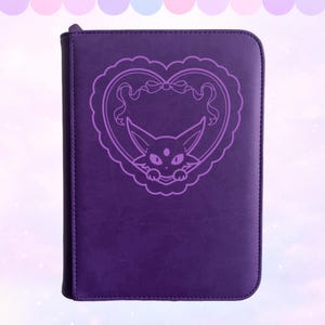 Puede incluir: Una carpeta morada oscura con cremallera con un diseño en forma de corazón lila claro con un gato de dibujos animados. El diseño incluye un lazo y bordes festoneados. La carpeta es rectangular y tiene una textura suave.