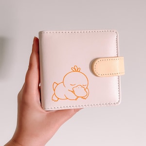 May include: A light pink square wallet featuring a small orange line art illustration of a sleeping duck-like creature on the front. The wallet includes a matching light pink body with a pale yellow snap closure tab and visible stitching.