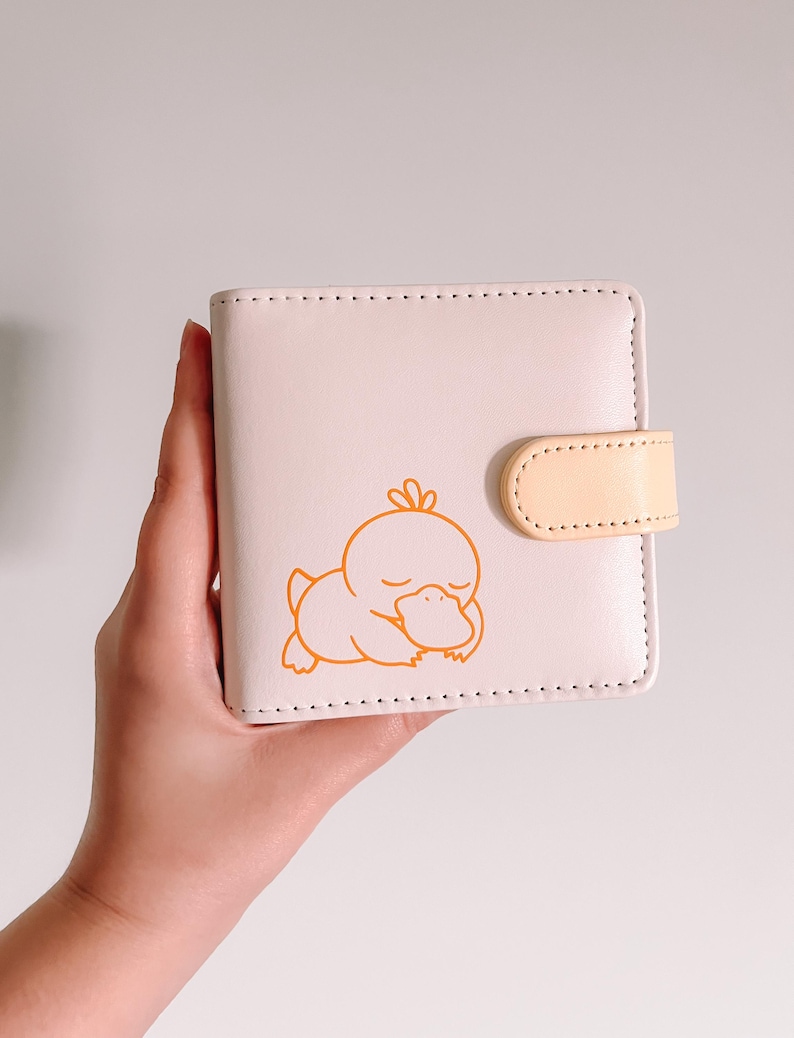May include: A light pink square wallet featuring a small orange line art illustration of a sleeping duck-like creature on the front. The wallet includes a matching light pink body with a pale yellow snap closure tab and visible stitching.
