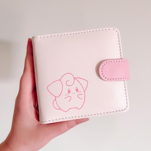 May include: A light pink bi-fold wallet featuring a darker pink line art illustration of a round, pointed-eared creature on the front cover. The wallet includes a matching pink snap-button closure tab on the right side with visible stitching.