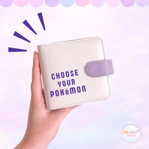 May include: A square, off-white bi-fold wallet featuring a purple snap closure and the text "CHOOSE YOUR POKEMON" printed in purple on the front. The wallet has visible stitching along the edges and a smooth, matte finish on the exterior surface.