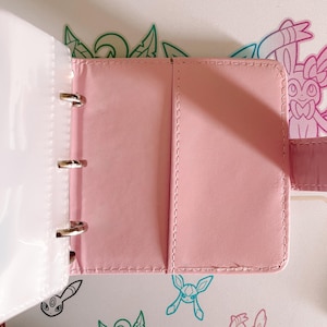 May include: A light pink binder with a smooth finish and stitched edges, featuring a three-ring metal mechanism on the left side and a vertical pocket on the right interior panel. The binder is open, revealing a clean, solid-colored surface.