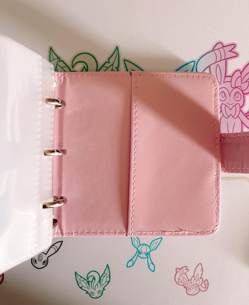 May include: A light pink binder with a smooth finish and stitched edges, featuring a three-ring metal mechanism on the left side and a vertical pocket on the right interior panel. The binder is open, revealing a clean, solid-colored surface.