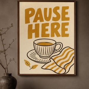 May include: A framed wall art print featuring a cream background with the mustard yellow text PAUSE HERE. Below the text is a line-drawn illustration of a teacup on a saucer, two small leaves, and a folded cloth with mustard yellow stripes.