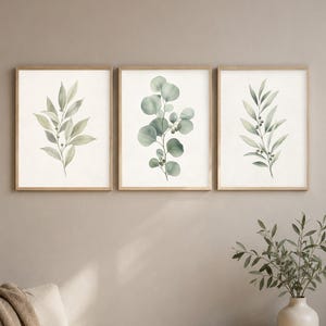 Può includere: Un set di tre stampe artistiche rettangolari da parete con cornici in legno chiaro. Ogni stampa presenta un'illustrazione ad acquerello di fogliame botanico verde su sfondo bianco, raffigurante varie forme di foglie e piccole bacche nei toni del verde.