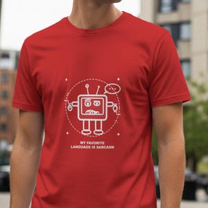 May include: A red t-shirt featuring a white cartoon robot design with the text "MY FAVORITE LANGUAGE IS SARCASM". The robot has a square head, antennas, and a speech bubble. The shirt is made of a soft material.