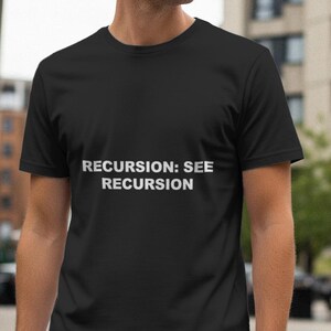 May include: Black t-shirt with the white text "RECURSION: SEE RECURSION". The t-shirt is a crew neck style. The background shows buildings and a street scene.
