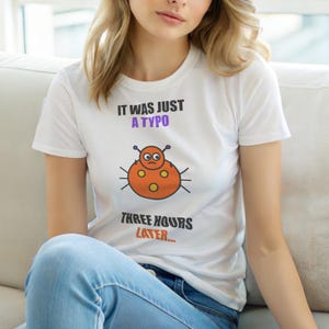 Może przedstawiać: Biała koszulka z napisem "IT WAS JUST A TYPO" w kolorze fioletowym i grafiką z kreskówkowym robakiem. Poniżej grafiki napis "THREE HOURS LATER..." w kolorze pomarańczowym. Koszulka jest z krótkim rękawem.