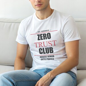 Op de afbeelding: Wit T-shirt met de tekst "ZERO TRUST CLUB" in zwart en rood, met "ACCESS DENIED UNTIL PROVEN" eronder. Het shirt heeft ook de tekst "- EST. PARANOIA -" boven de hoofdtekst. De persoon draagt een blauwe spijkerbroek.