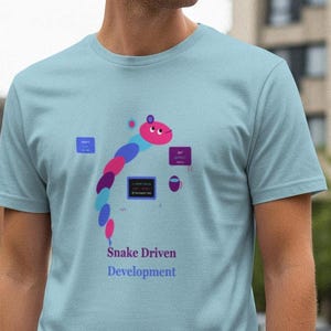 Op de afbeelding: Lichtblauw t-shirt met een kleurrijke slangengrafiek en de tekst "Snake Driven Development". Het slangontwerp bevat roze, blauwe en paarse vormen. Het shirt heeft een ronde hals en korte mouwen.