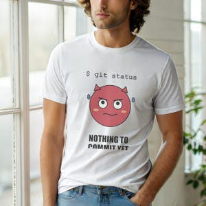 May include: White t-shirt featuring a red devil emoji with the text "$ git status" and "NOTHING TO COMMIT YET". The short-sleeved shirt has a round neck.
