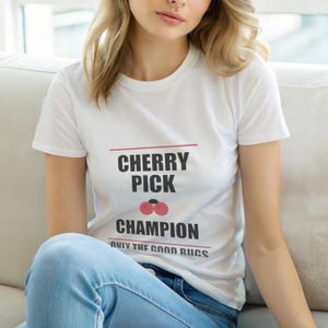 Puede incluir: Camiseta blanca con el texto "CHERRY PICK CHAMPION" en negro, con una línea roja encima y debajo del texto. La camiseta también presenta un gráfico de cereza y las palabras "ONLY THE GOOD RUCS" en negro.