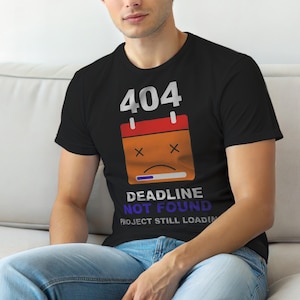 Może przedstawiać: Czarny t-shirt z grafiką przedstawiającą numer "404" nad ikoną kalendarza z nieszczęśliwą twarzą i napisem "DEADLINE NOT FOUND PROJECT STILL LOADING". Koszulka jest noszona przez osobę.