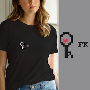 May include: A black short-sleeved t-shirt featuring a small pixel art graphic on the left chest. The graphic consists of a grey key icon with a red heart shape in the centre, followed by the black text FK in a pixelated font style.
