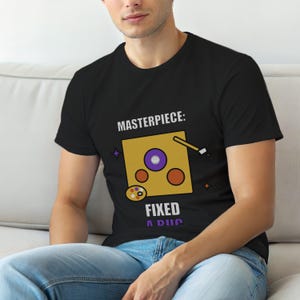 Puede incluir: Camiseta negra con el texto "MASTERPIECE: FIXED A DUC" sobre un gráfico de un cuadrado amarillo con un círculo morado, círculos naranjas, un pincel y una paleta. La camiseta la lleva una persona.