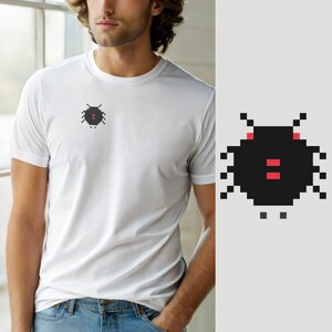 Op de afbeelding: Een wit T-shirt met korte mouwen en een kleine pixel art afbeelding op de linkerborst. De afbeelding toont een zwart, rond, insectachtig wezen met twee rode ogen en twee rode rechthoekige markeringen in het midden van het lichaam.