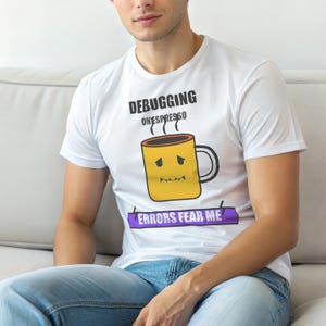 Peut inclure: T-shirt blanc avec l'inscription "DEBUGGING ONESPRESSO" au-dessus d'un dessin de tasse à café jaune avec un visage triste. En dessous, une bannière violette indique "ERRORS FEAR ME".