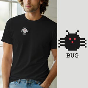 Op de afbeelding: Een zwart T-shirt met korte mouwen en een kleine, gepixelde afbeelding op de linkerborst. De afbeelding toont een zwart, insectachtig wezen met drie rode, vierkante ogen en het woord BUG in een eenvoudig schreeflettertype eronder.