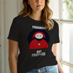 May include: Black t-shirt with the words "PARANOID" and "BUT PREPARED" in white. The shirt features a graphic of a red figure with a lock symbol. The figure is inside a dark blue circle.