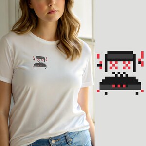 Może przedstawiać: Biały t-shirt z krótkim rękawem z małą grafiką pixel art na lewej piersi. Wzór przedstawia ciemnoszarą postać robota z czerwonymi, pikselowymi oczami, czerwonymi akcentami po bokach oraz czarną podstawą z trzema małymi czerwonymi kwadratami.