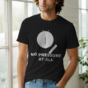 Może przedstawiać: Czarny t-shirt z białym nadrukiem bębna i pałeczki oraz napisem "NO PRESSURE AT ALL". Koszulka ma okrągły dekolt.