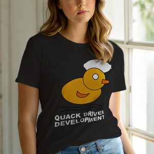 Op de afbeelding: Zwart T-shirt met een cartoonontwerp van een gele eend en een witte wolk. De tekst "QUACK DRIVEN DEVELOPMENT" staat eronder gedrukt. Het T-shirt is gemaakt van een zacht materiaal. Het ontwerp is een humoristische kijk op softwareontwikkeling.