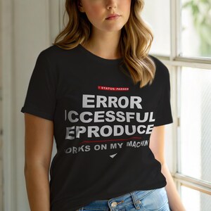 Op de afbeelding: Zwart T-shirt met witte tekst: "ERROR SUCCESSFUL REPRODUCE WORKS ON MY MACHIN". Rode tekst erboven luidt "STATUS: PASSED". Het T-shirt heeft een ronde hals.