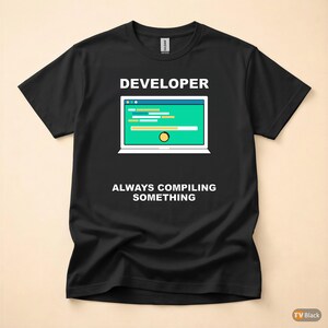 Op de afbeelding: Zwart t-shirt met het woord "DEVELOPER" boven een laptopafbeelding. Het laptopscherm toont code en een voortgangsbalk. De tekst "ALWAYS COMPILING SOMETHING" staat eronder.