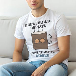 May include: White t-shirt featuring the text "BREW. BUILD. DEPLOY." above a brown coffee mug graphic with an infinity symbol and the text "REPEAT UNTIL STABLE" below. The shirt is worn by a person.