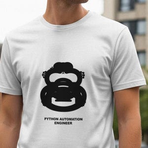 Op de afbeelding: Wit T-shirt met een zwart grafisch ontwerp van een gestileerd object en de tekst "PYTHON AUTOMATION ENGINEER". Het shirt heeft korte mouwen en een ronde hals.
