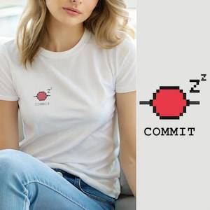Puede incluir: Una camiseta blanca de manga corta con un pequeño gráfico de pixel art en el pecho. El diseño muestra un círculo rojo con contornos negros, un pequeño símbolo de z al cuadrado encima y la palabra COMMIT en letras mayúsculas negras.