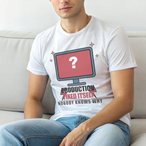May include: White t-shirt with a computer monitor graphic displaying a question mark. The text below reads "PRODUCTION FIXED ITSELF, NOBODY KNOWS WHY". The design includes star accents.