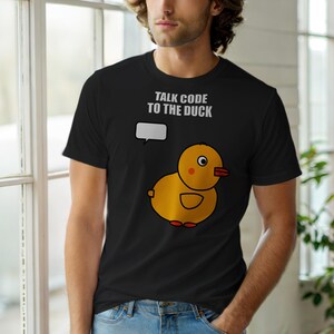 May include: Black t-shirt featuring a cartoon yellow duck with orange accents and the text "TALK CODE TO THE DUCK" in white. The duck has a speech bubble above it. The shirt is made of a soft material.