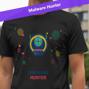 Puede incluir: Camiseta negra con un diseño gráfico colorido y el texto "MALWARE HUNTER" en rojo y azul. El diseño incluye un símbolo de diana y varias formas coloridas. El texto "Malware Hunter" también está impreso en una pancarta morada.