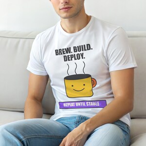 Op de afbeelding: Wit T-shirt met de tekst "BREW. BUILD. DEPLOY." boven een lachende gele koffiemok. Daaronder staat "REPEAT UNTIL STABLE" op een paarse banner. De persoon draagt een blauwe spijkerbroek.
