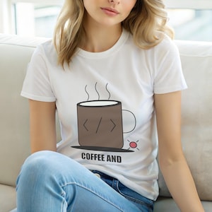 Op de afbeelding: Wit t-shirt met een bruine koffiemok afbeelding, code symbolen en stoom. De tekst "COFFEE AND" staat eronder gedrukt. De persoon draagt een blauwe spijkerbroek.