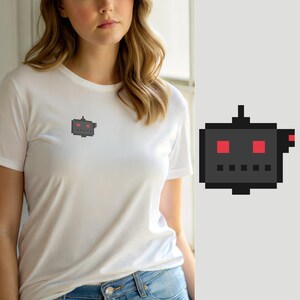 May include: A white short-sleeved t-shirt featuring a small pixel art graphic on the left chest. The graphic depicts a dark gray square robot head with two bright red square eyes, a small red side detail, and a row of five small dark gray squares.