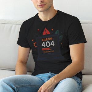 May include: Black t-shirt with a graphic of a laptop screen showing an "ERROR 404" message, a red warning symbol, and the word "Rebooting". The design includes orange and green details.