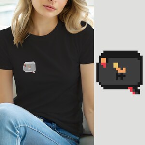 Puede incluir: Una camiseta negra de manga corta con un pequeño gráfico de pixel art en el pecho izquierdo. El gráfico es un bocadillo de diálogo gris oscuro que contiene un personaje pixelado naranja y amarillo con un pequeño acento rojo en la esquina.