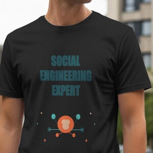 May include: Black t-shirt with the text "SOCIAL ENGINEERING EXPERT" in teal. Below the text is a graphic design with orange and teal circles and lines. The shirt is made of a soft material.