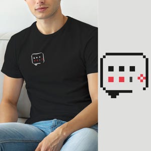 Puede incluir: Una camiseta negra de manga corta con un pequeño gráfico de pixel art en el pecho izquierdo. El gráfico es el contorno de un bocadillo de diálogo gris claro que contiene tres cuadrados negros, dos cuadrados rojos, un pequeño rectángulo gris y una cruz roja.