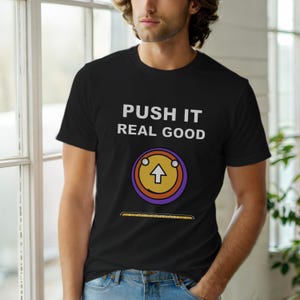 Op de afbeelding: Zwart t-shirt met de witte tekst "PUSH IT REAL GOOD". Het heeft een afbeelding van een gele cirkel met een pijl omhoog, omringd door paarse en oranje ringen. Het t-shirt heeft een ronde hals.
