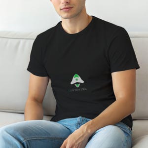 Op de afbeelding: Zwart t-shirt met een wit en groen grafisch ontwerp. Het ontwerp heeft een gestileerde pijlvorm met de tekst "$SHIPPING" eronder. Het shirt is gemaakt van een zacht materiaal.